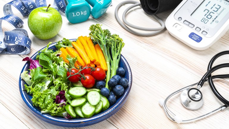 Eine Schale mit Gemüse, ein Apfel, Hanteln und ein Blutdruckmessgerät