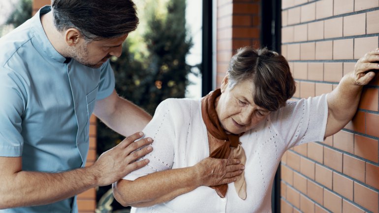 Eine ältere Dame lehnt an einer Wand und hält Ihre Hand auf ihr Herz